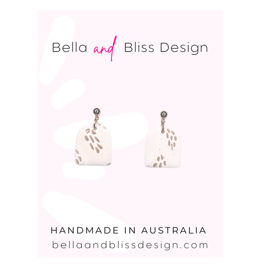 White earrings with black speckles on a pink and white gradient background with 'Bella and Bliss Design' branding.