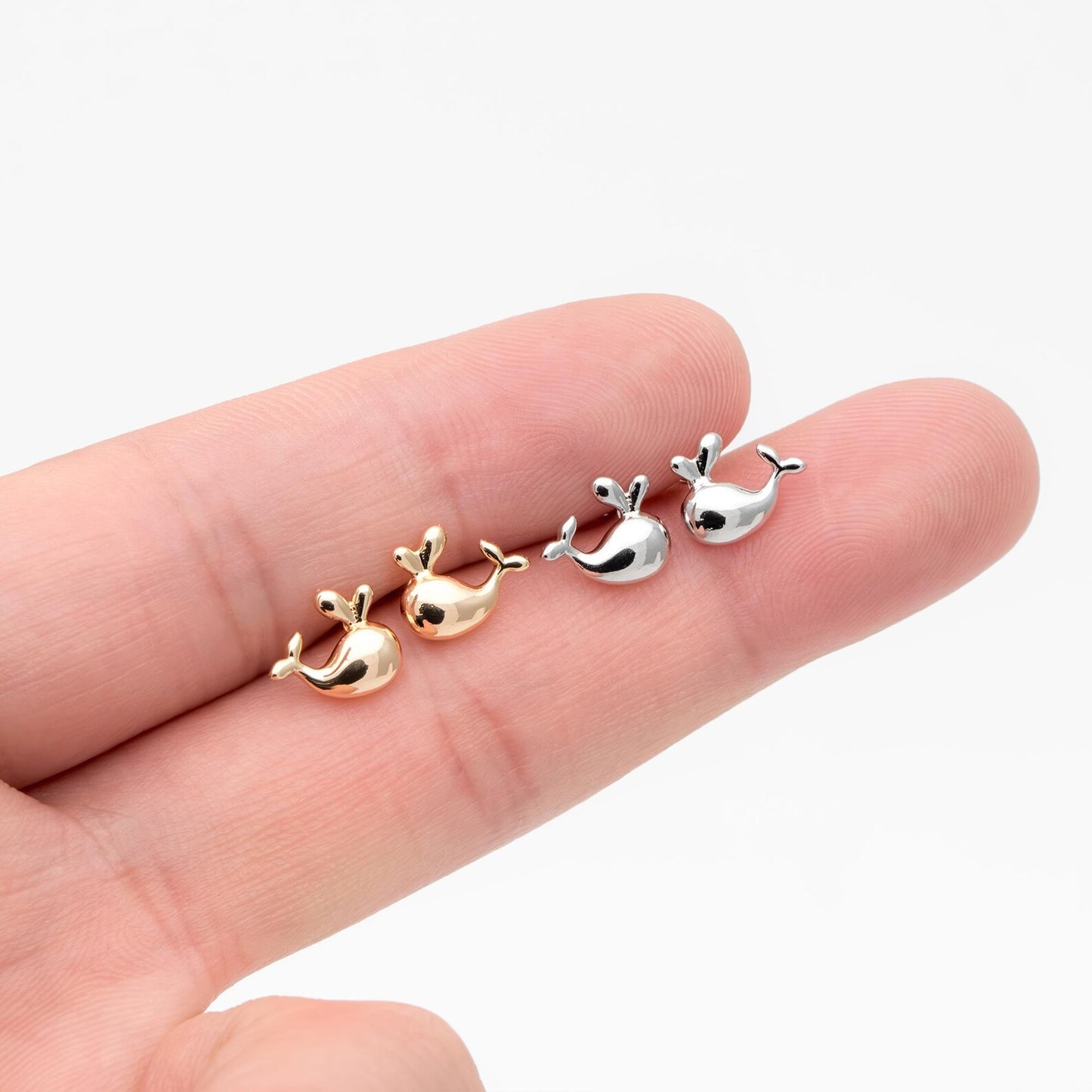 Four small whale-shaped earrings in gold, rose gold, and silver held between fingers on a white background.