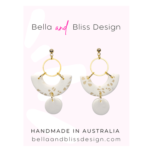 White earrings with gold accents on a pink background, featuring Bella and Bliss Design branding.