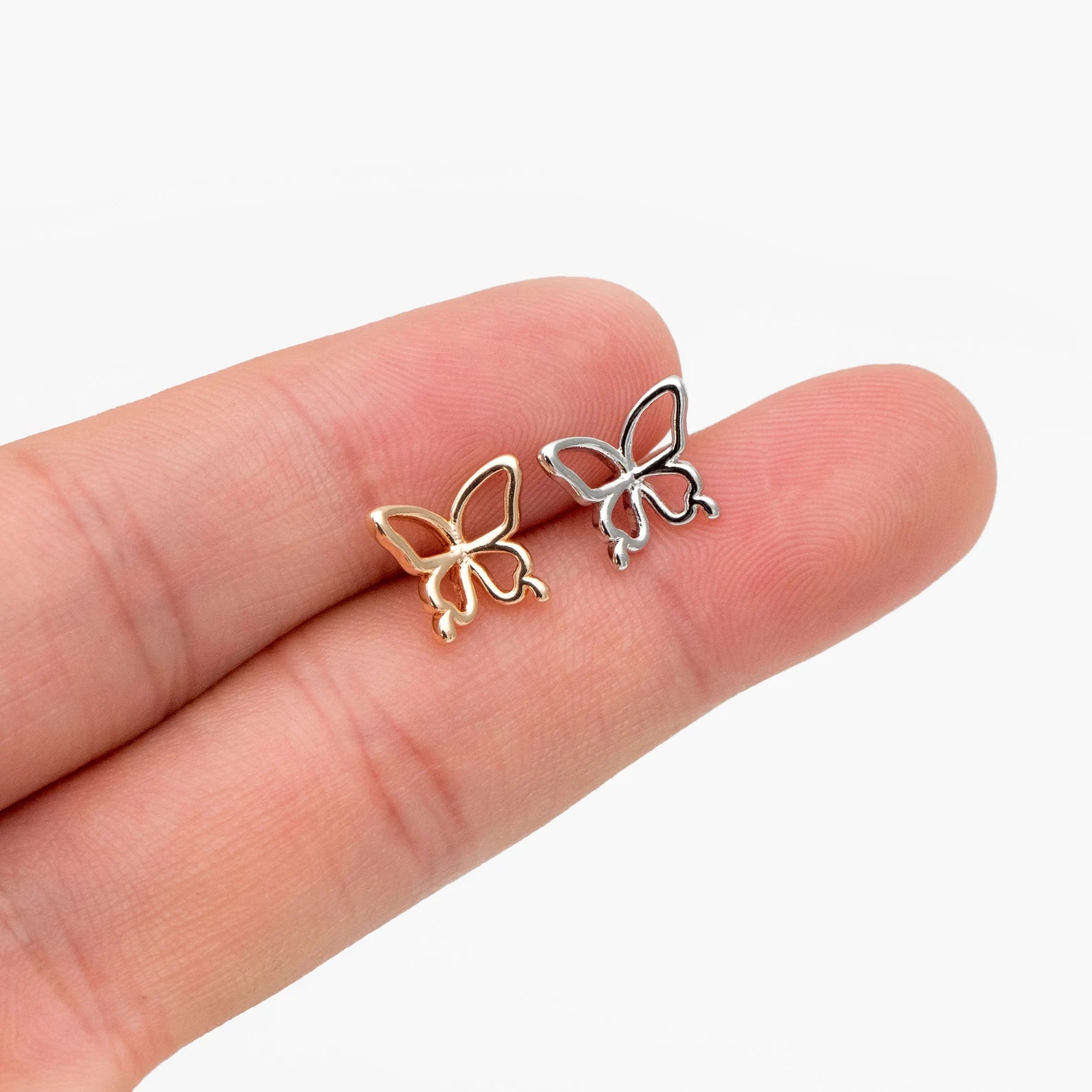 Butterfly-shaped earrings in gold and silver held between fingers on a white background