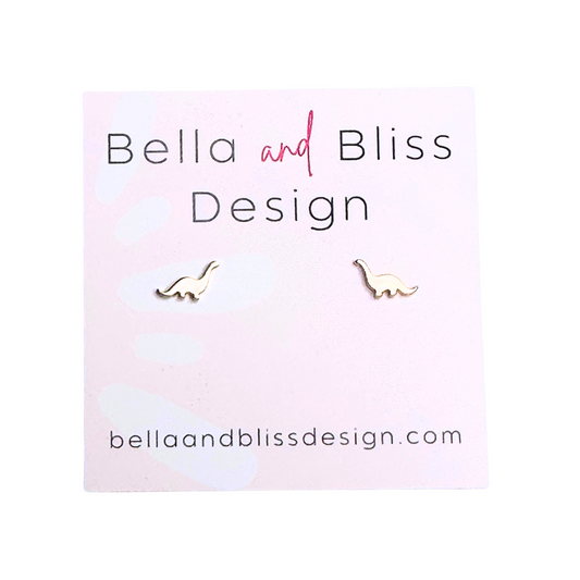 A pair of gold mini stud earrings with a dinosaur design, displayed on a pink background with the brand name 'Bella and Bliss Design'.