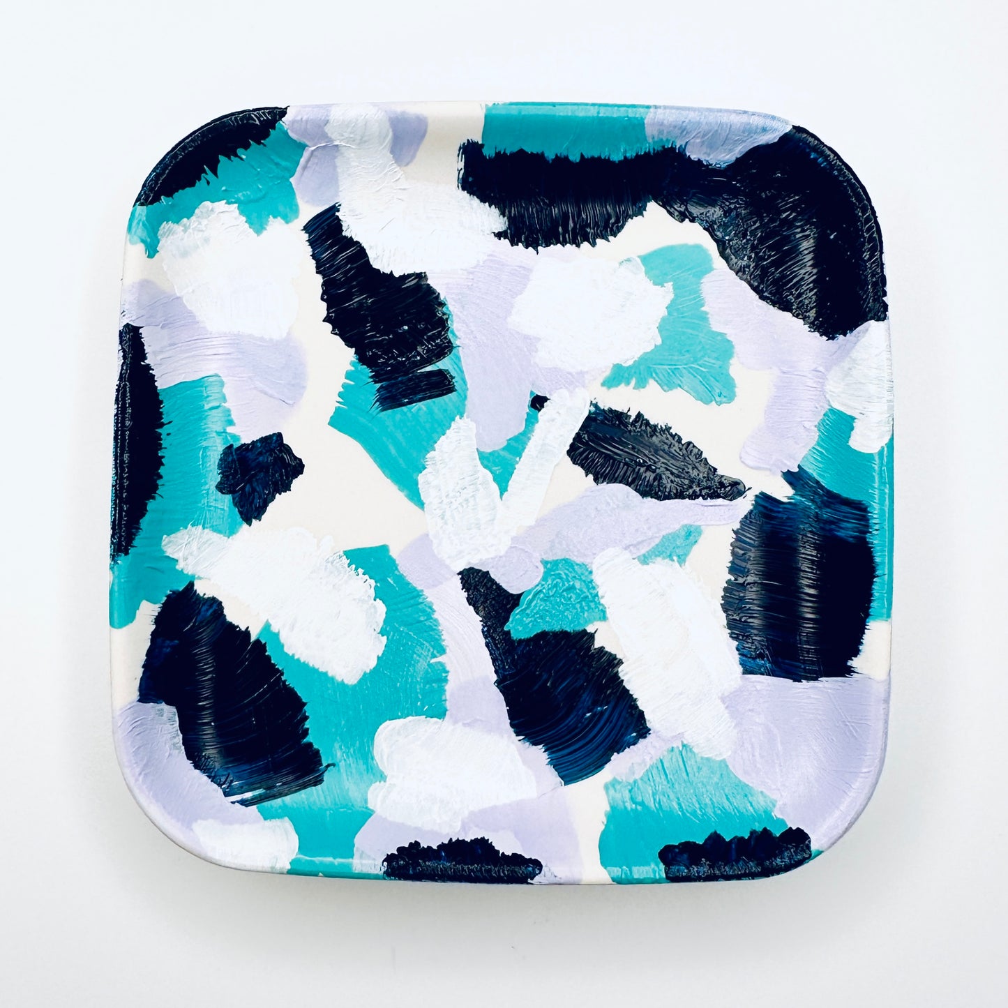 A hand-painted jewellery dish featuring a geometric pattern in shades of deep blue, light jade, lilac, and white.